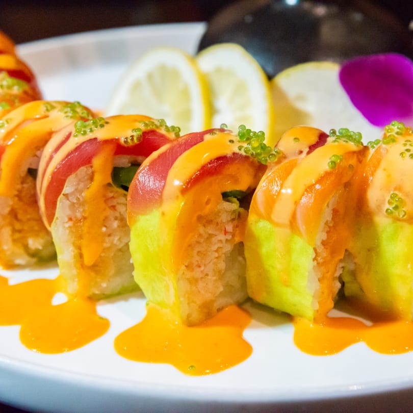 Close-up of sushi rolls with avocado, tuna, orange sauce, and green roe, garnished with lemon slices.