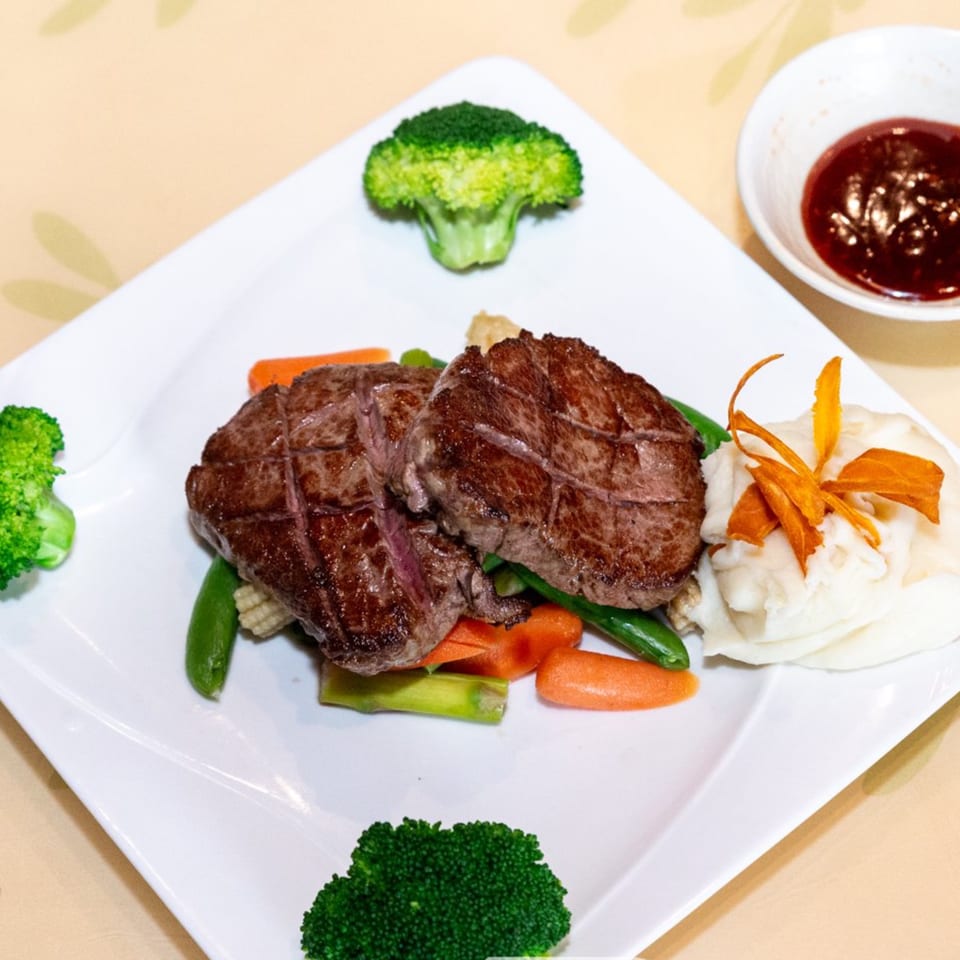 Two grilled steak medallions with mixed vegetables, broccoli, and a side of sauce on a white plate.